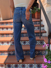 A person viewed from behind wearing medium-wash blue jeans with a straight-leg or slightly flared fit, standing on outdoor tiled stairs with plants and a black railing visible.