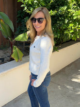 A woman wearing a white ribbed knit long-sleeve sweater with a small black embroidered polo player logo on the chest, paired with blue jeans and black sunglasses, standing outdoors on a concrete walkway with green foliage and a beige wall behind her.