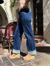 A person wearing dark blue wide-leg cuffed jeans with light blue contrast stitching, paired with tan wedge sandals, standing on a stone patio in front of a building with red-and-white patterned chairs.