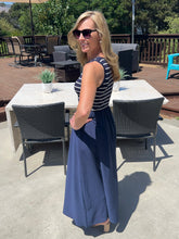 Woman in a blue dress standing outdoors on a patio with tables and chairs.