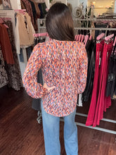 A woman viewed from behind wearing a long-sleeve button-up blouse with a vibrant orange, pink, and blue geometric pattern, paired with light blue jeans in a clothing boutique.