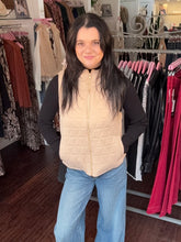 A woman standing in a clothing boutique wearing a quilted beige puffer vest over a black long-sleeve top and light blue jeans.
