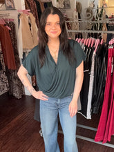 A woman with long dark hair stands in a clothing boutique wearing a dark green draped short-sleeve top and light blue jeans, with racks of clothes visible in the background.