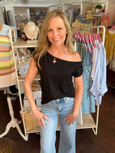 Woman standing wearing a black top off one shoulder in a clothing store with racks of clothes in the background