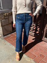 A person wearing dark blue cropped jeans with contrast stitching, a cream ribbed knit sweater, and tan slide sandals, standing outdoors on a tiled porch.