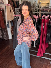 A woman with long dark hair stands in a clothing boutique wearing a long-sleeve V-neck blouse with a multicolor abstract print in orange, blue, and red, paired with light-wash blue jeans.