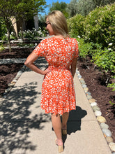 Woman in an orange floral dress standing on a garden path with greenery around.