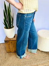 Person wearing blue jeans standing next to a plant and wooden box on a textured floor.