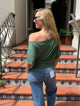 A woman wearing a green off-the-shoulder top and blue jeans stands on outdoor tiled steps, viewed from behind. The product being shown is the green top, with a white tag still attached near the back pocket.