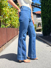 A person wearing blue high-waisted flare jeans with a white top and beige sandals, standing outdoors on a paved path with greenery and a house in the background.
