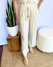 Person wearing beige striped pants standing next to a plant and wooden box on a textured floor.