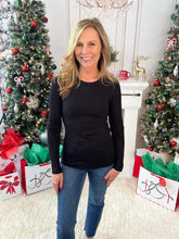 Woman wearing a black long sleeve crew neck t-shirt standing in a festive room with Christmas trees and decorations.