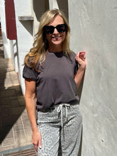 A woman wearing a dark gray short-sleeve t-shirt and gray leopard-print sweatpants, standing outdoors against a textured light gray wall.