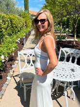 Woman in a white dress standing outdoors with greenery and a bench in the background