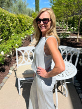 Woman in a light gray sleeveless dress standing outdoors with greenery and a white chair in the background.