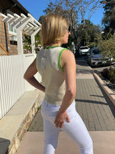 Woman in a sleeveless top and white pants standing on a sidewalk with a house and cars in the background.