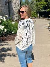 Woman wearing a white open-knit cardigan and blue jeans standing on a sidewalk with greenery in the background.