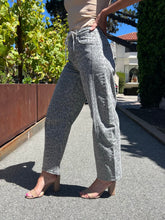 A person outdoors wearing light gray wide-leg jeans with a subtle leopard print pattern, paired with beige heeled sandals and a beige top. The background shows a sunny residential street with greenery, a brown fence, and a house.