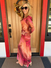 A woman wearing a long-sleeve maxi dress with a pink, orange, and red abstract swirl pattern, standing in front of a wooden front door with red trim.