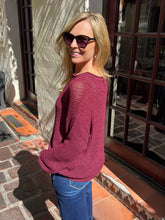 A woman wearing a maroon knit long-sleeve sweater and blue jeans, standing outdoors in front of a building with glass doors and tiled floors.