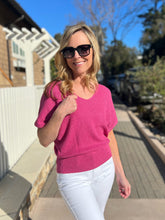 Woman in a magenta sweater and white pants standing outdoors on a sunny day.