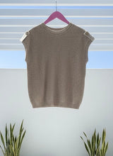 Khaki knit sleeveless sweater on a hanger against a light background with plants at the bottom.