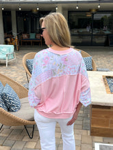 Woman in a pink patchwork top and white pants standing outdoors near patio furniture.