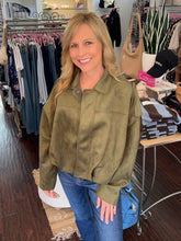 A woman with blonde hair smiling in a boutique, wearing an olive green cropped suede jacket and blue jeans.