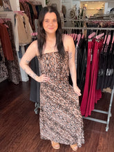 A woman modeling a strapless snake-print maxi dress in a clothing boutique, standing in front of racks of other garments.