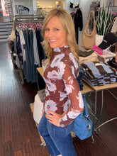 A woman posing in a clothing boutique wearing a sheer brown mock-neck long-sleeve top with a white and pink floral pattern, paired with blue jeans.