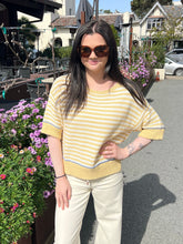 Woman wearing a yellow striped sweater and sunglasses standing outdoors with flowers and buildings in the background