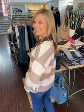 A woman with blonde hair smiling and looking over her shoulder inside a clothing boutique. She is wearing a chunky knit sweater with large beige and white stripes, paired with blue jeans.