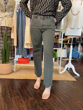 A person standing in a clothing boutique wearing straight-leg gray jeans, a black-and-white patterned long-sleeve top, and clear-strap heeled sandals. Clothing racks, mannequins, a cactus plant, and wood flooring are visible in the background.