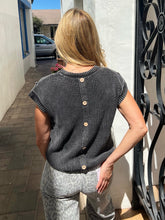 A woman viewed from behind wearing a dark gray ribbed knit short-sleeve top with three light wood buttons down the back, paired with light gray patterned pants, standing outdoors next to a white wall and black wrought iron gate.