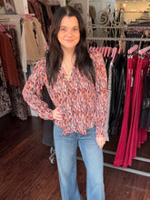 A woman with long dark hair stands in a clothing boutique wearing a long-sleeve V-neck blouse with a multicolor abstract print in orange, blue, and red, paired with light-wash blue jeans.