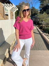 Woman in a magenta sweater and white pants standing outdoors on a sunny day.