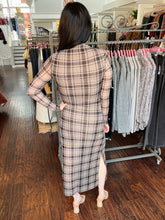 A woman viewed from behind wearing a beige and brown plaid long-sleeve midi dress with a side slit, standing in a clothing boutique next to racks of other garments.