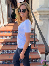 A woman wearing a white short-sleeve crewneck t-shirt and dark jeans stands on outdoor terracotta tiled steps wearing black sunglasses.