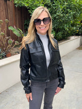A woman wearing a black faux leather moto jacket, gray pants, and a gray turtleneck stands outdoors in a backyard setting with green plants and a wooden fence.