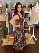 A woman modeling a brown sleeveless maxi dress with a blue-gray floral pattern, standing inside a boutique clothing store.