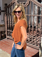 A woman wearing a rust-orange short-sleeve knit top and blue jeans stands outdoors in front of a decorative brown metal gate.