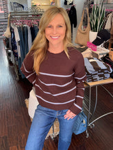 A smiling blonde woman wearing a brown crewneck sweater with white horizontal stripes and blue jeans, standing inside a clothing boutique.