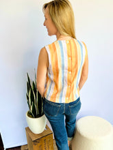 Woman wearing a striped sleeveless top and jeans standing in a room with a plant and ottoman.