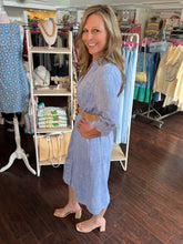 Woman in a blue dress standing in a clothing store with racks of clothes and accessories.