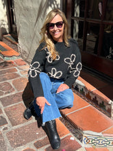 A woman wearing a black knit sweater with white floral patterns, blue jeans, and black ankle boots sits on a brick ledge outdoors.