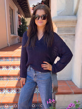A woman wearing a dark navy blue knit V-neck sweater and blue jeans, standing on outdoor tiled steps in front of a building.