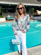 Woman standing by a pool wearing a patterned blouse and white pants, holding a light blue handbag.