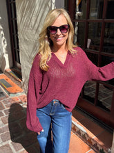 A woman wearing a maroon knit V-neck sweater and blue jeans, standing outdoors in front of a building with glass doors.