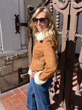 A woman wearing a tan suede moto jacket with white shearling-style trim, blue jeans, and sunglasses, standing outdoors next to a decorative metal gate.
