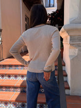 A woman viewed from behind wearing a cream ribbed knit sweater with black trim details on the collar and cuffs, paired with blue jeans, standing on terracotta tiled steps.